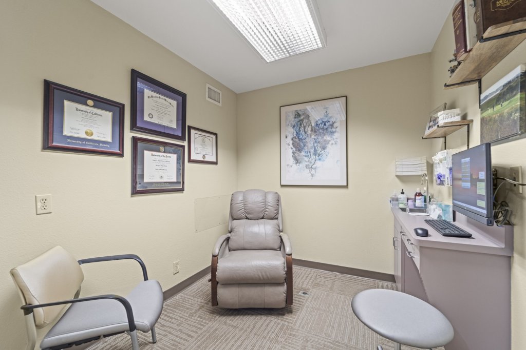 Private consultation room: beige reclining dental chair, exam stool, computer workstation, framed certificates on wall, and abstract artwork