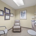 Private consultation room: beige reclining dental chair, exam stool, computer workstation, framed certificates on wall, and abstract artwork
