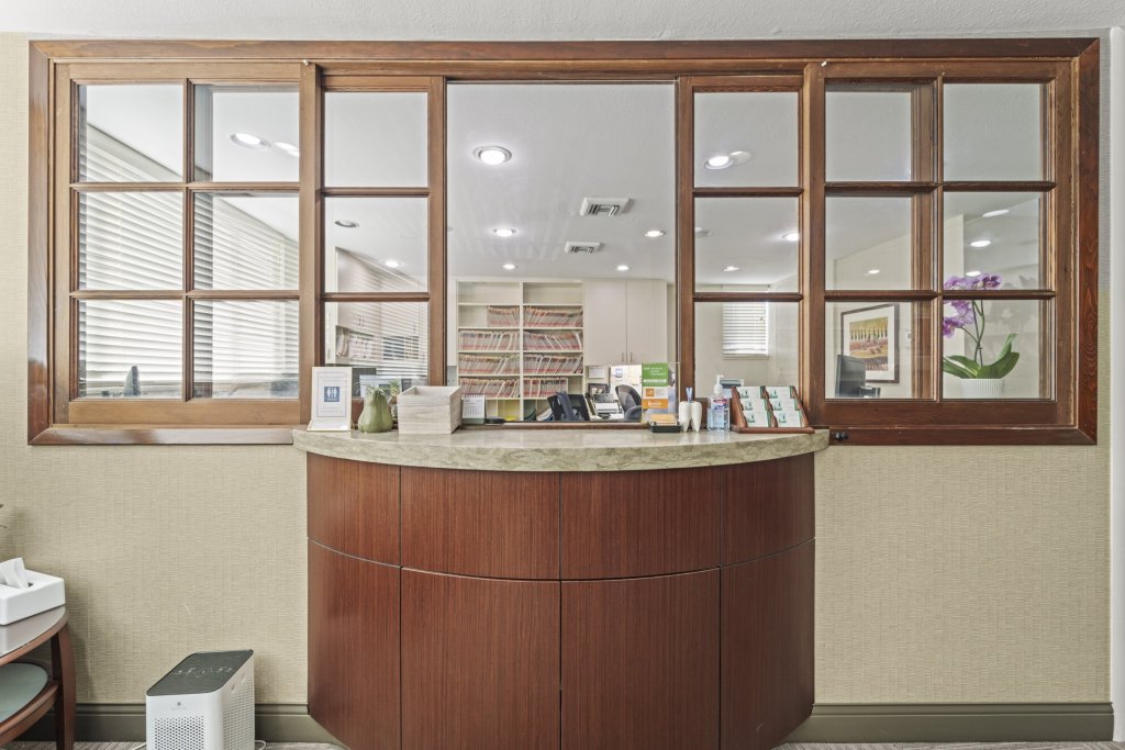Front desk with curved wood cabinetry and stone countertop; behind it, a glass-paneled partition reveals the clinical area with cabinets and supplies