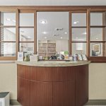 Front desk with curved wood cabinetry and stone countertop; behind it, a glass-paneled partition reveals the clinical area with cabinets and supplies