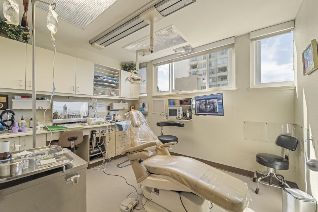 Well-lit dental treatment room: beige patient chair, overhead surgical light, instrument tray, sterilization unit, wall-mounted monitor, and large windows providing natural light