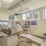 Well-lit dental treatment room: beige patient chair, overhead surgical light, instrument tray, sterilization unit, wall-mounted monitor, and large windows providing natural light