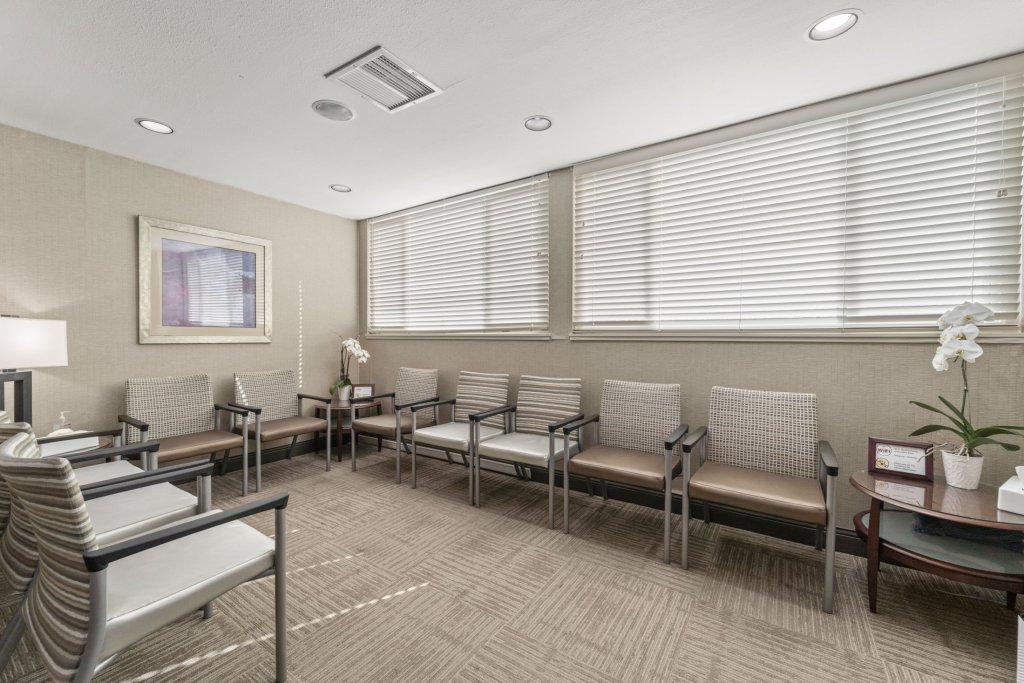Spacious waiting area with eight modern armchairs arranged in rows, neutral carpeting, large windows with blinds, wall art, and side tables with plants