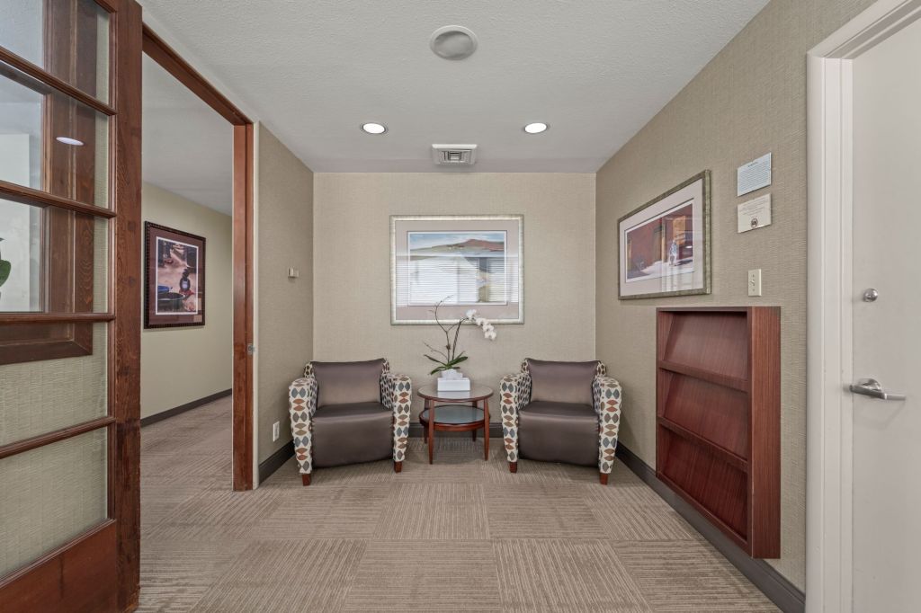 Entry hallway with open wooden doorframe, two upholstered armchairs, framed artwork, potted orchid, and built-in bookshelf with brochures