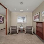 Entry hallway with open wooden doorframe, two upholstered armchairs, framed artwork, potted orchid, and built-in bookshelf with brochures