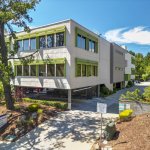 Modern dental office building with light gray stucco, green accent trim, large windows, and landscaped entrance walkway under clear blue sky