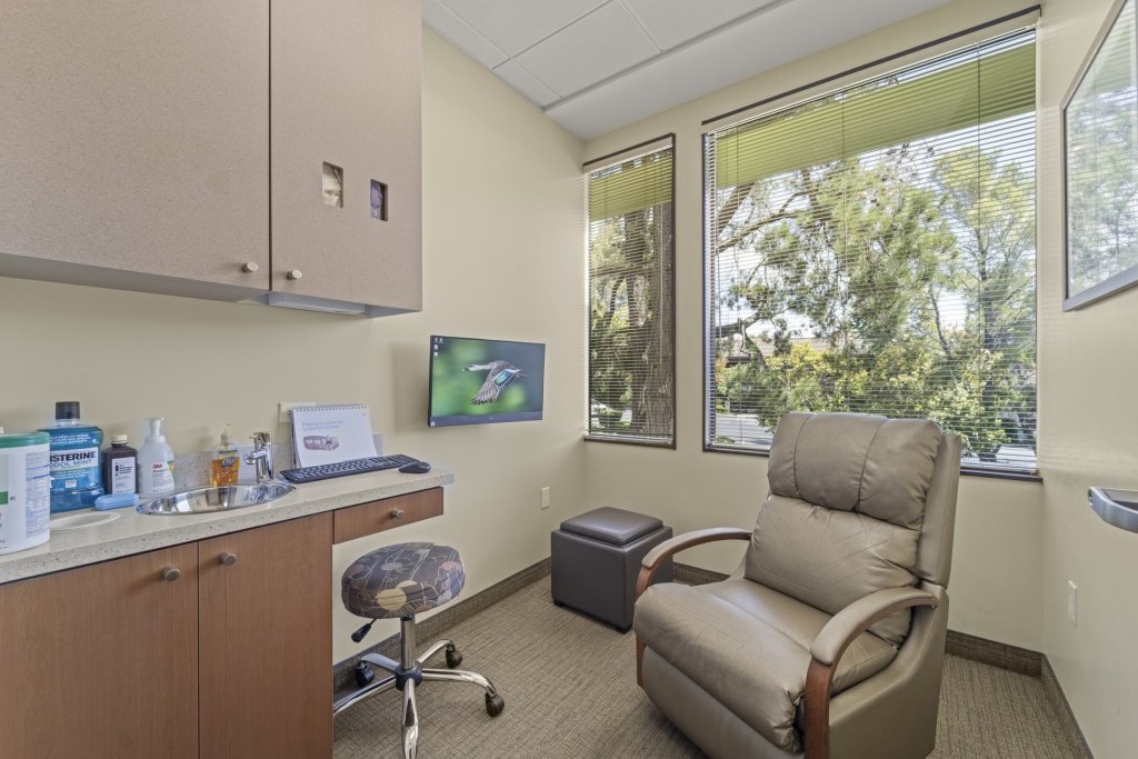 Private consultation/hygiene room: beige reclining chair, rolling stool, countertop with supplies, wall-mounted monitor, and large window with blinds and outdoor view