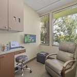 Private consultation/hygiene room: beige reclining chair, rolling stool, countertop with supplies, wall-mounted monitor, and large window with blinds and outdoor view