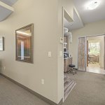 Interior hallway with neutral walls, recessed lighting, framed mirror/art, and open doorways revealing adjacent treatment rooms and a staff workstation
