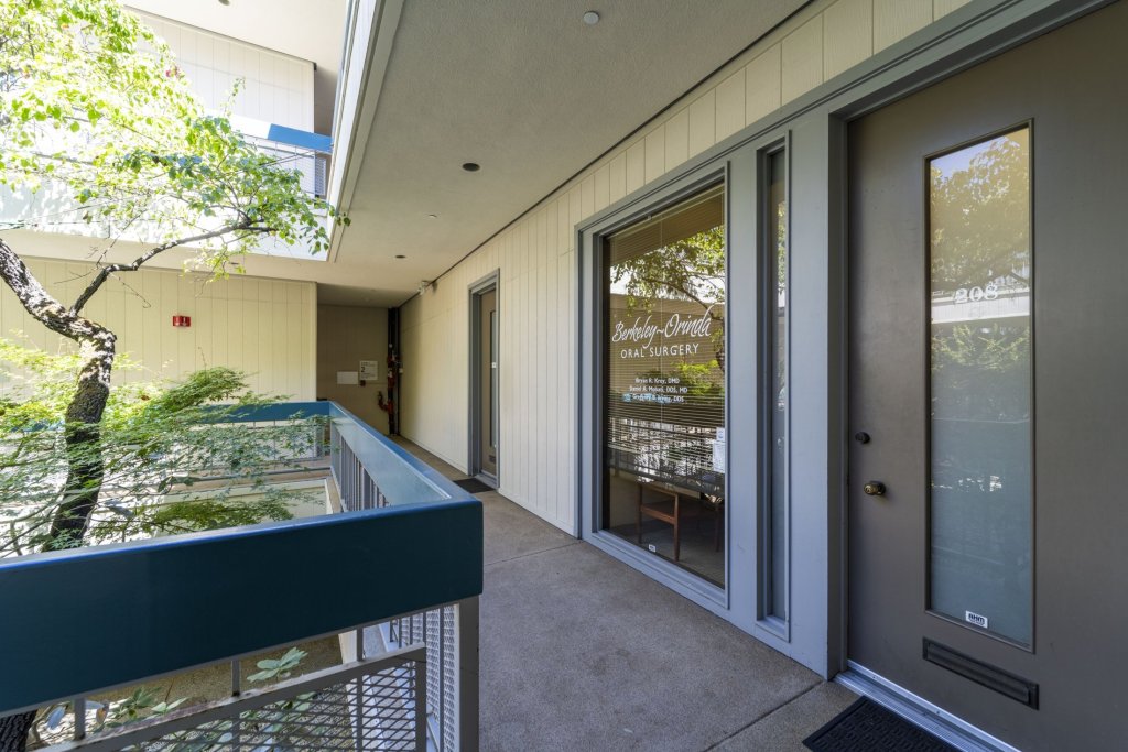 Exterior covered walkway with glass doors, metal railing, potted plants, and signage — leading to the main entrance of the dental practice