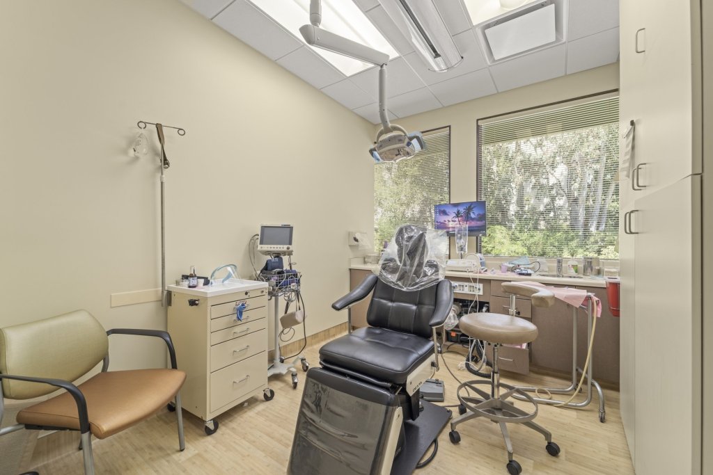 Primary treatment room: black dental chair with headrest, overhead lamp and delivery system, assistant’s stool, instrument cart, and skylight + window for natural illumination