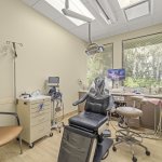 Primary treatment room: black dental chair with headrest, overhead lamp and delivery system, assistant’s stool, instrument cart, and skylight + window for natural illumination