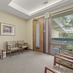 Calm waiting room: beige carpet, two floral-patterned armchairs, small side table with plant, framed artwork, and floor-to-ceiling glass door showing outdoor greenery