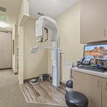 Digital radiography room: upright panoramic X-ray machine, counter with computer monitor displaying dental imagery, storage cabinets, and stool — dedicated for imaging diagnostics